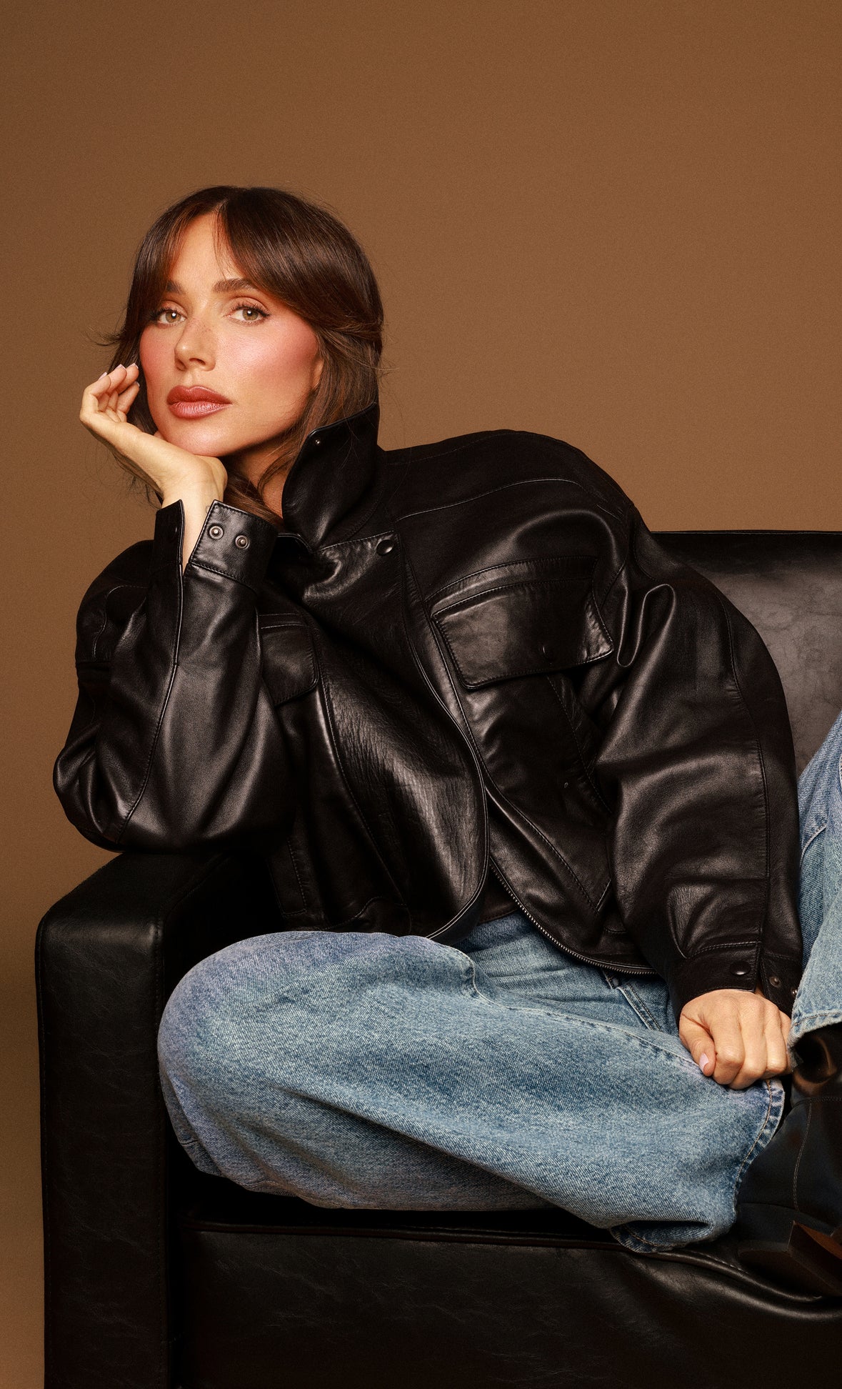Woman wearing a black leather jacket and blue jeans sitting on a brown background