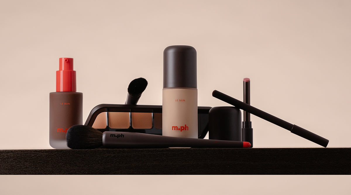 A collection of makeup products from the brand m.ph, including a liquid foundation bottle, a setting spray, an eyeshadow palette, brushes, and lip products, arranged on a dark surface against a warm beige background.
