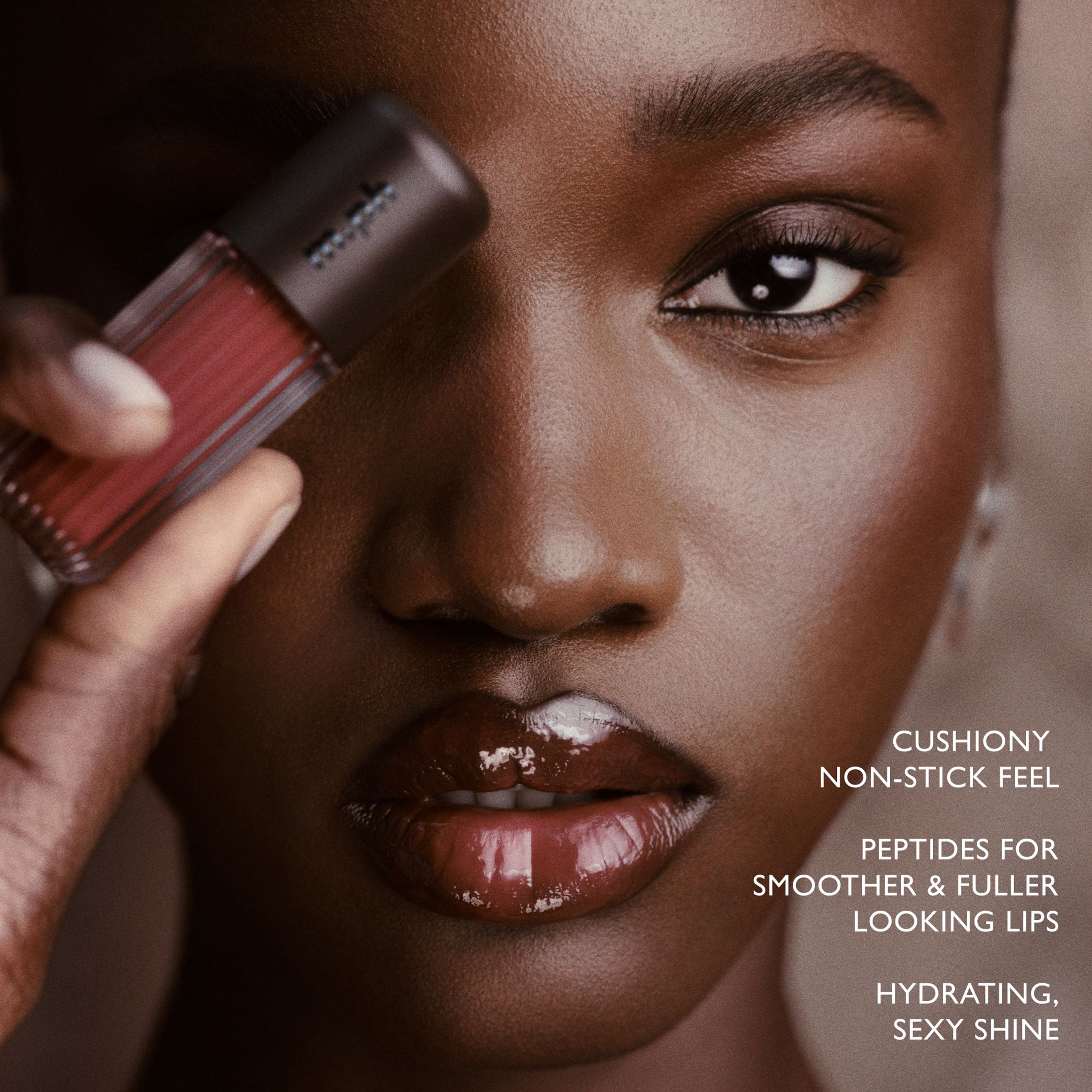 Close-up beauty shot of a model holding a lip gloss tube near her face, with glossy reddish-brown lips and promotional text on the right side.