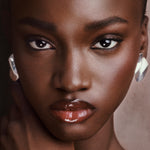 Close-up portrait of a model wearing large, chunky silver hoop earrings and glossy nude lips, set against a soft, warm neutral background.