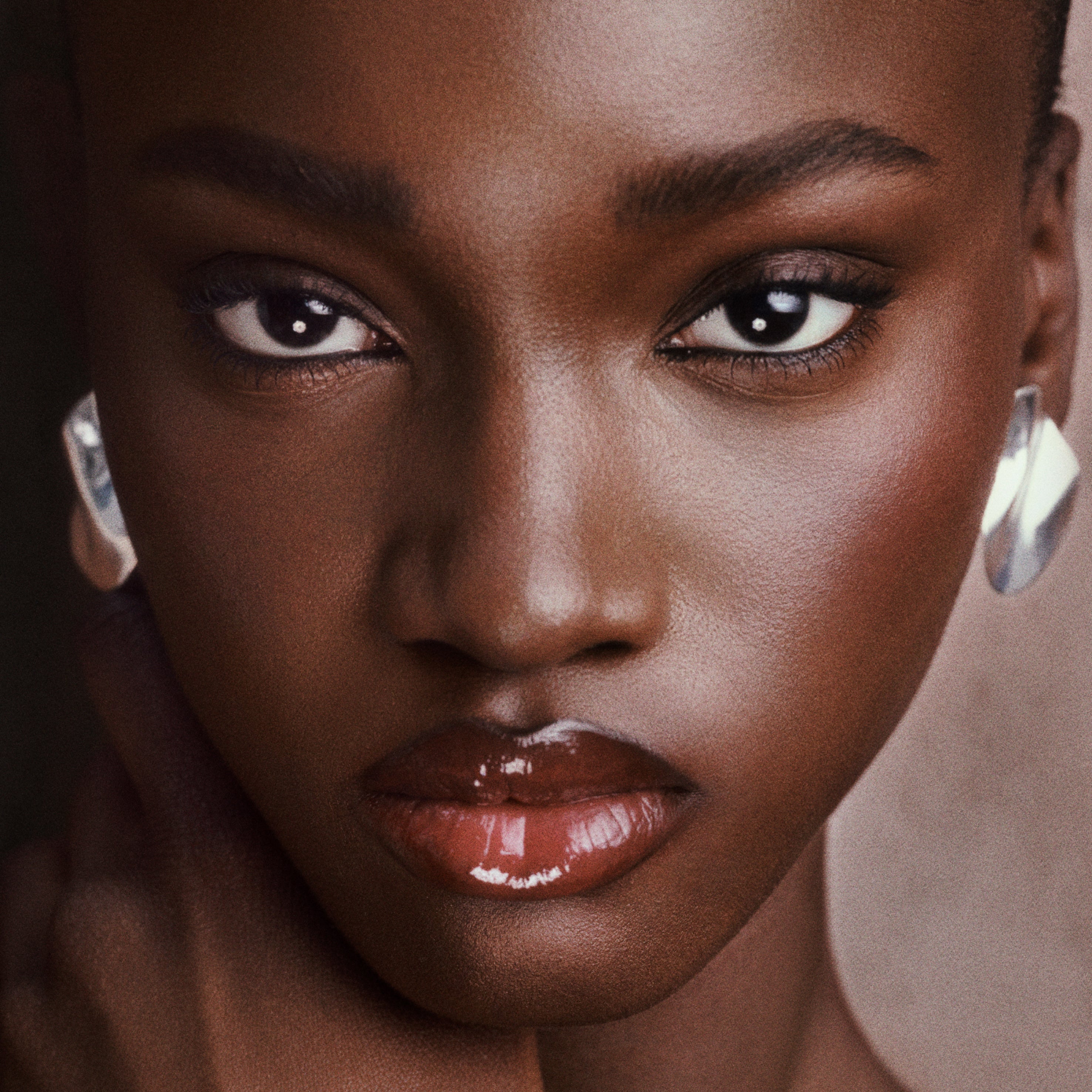 Close-up portrait of a model wearing large, chunky silver hoop earrings and glossy nude lips, set against a soft, warm neutral background.
