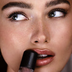 Close-up of a woman applying a dark lip product from a black tube with the brand name 'migh' visible, against a softly blurred warm-toned background.