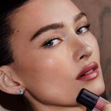 Close-up of a woman applying a pink lip product while wearing a sparkling stud earring. The lipstick tube is partially visible at the bottom right, and the background is a soft warm brown.