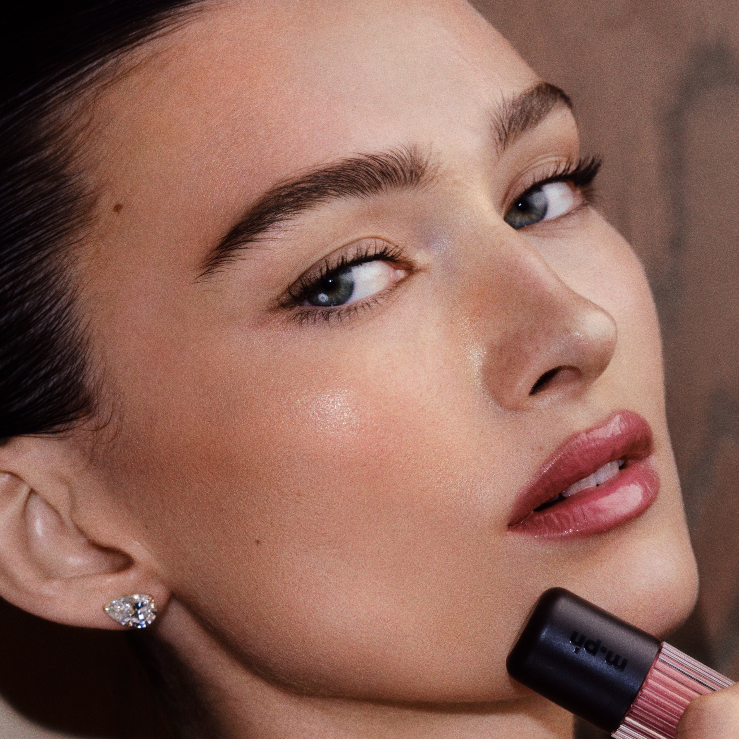 Close-up of a woman applying a pink lip product while wearing a sparkling stud earring. The lipstick tube is partially visible at the bottom right, and the background is a soft warm brown.