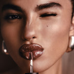 Close-up of a model applying glossy brown lip color with a doe-foot applicator, wearing large silver hoop earrings against a warm brown background.