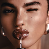 Close-up of a model applying glossy brown lip color with a doe-foot applicator, wearing large silver hoop earrings against a warm brown background.