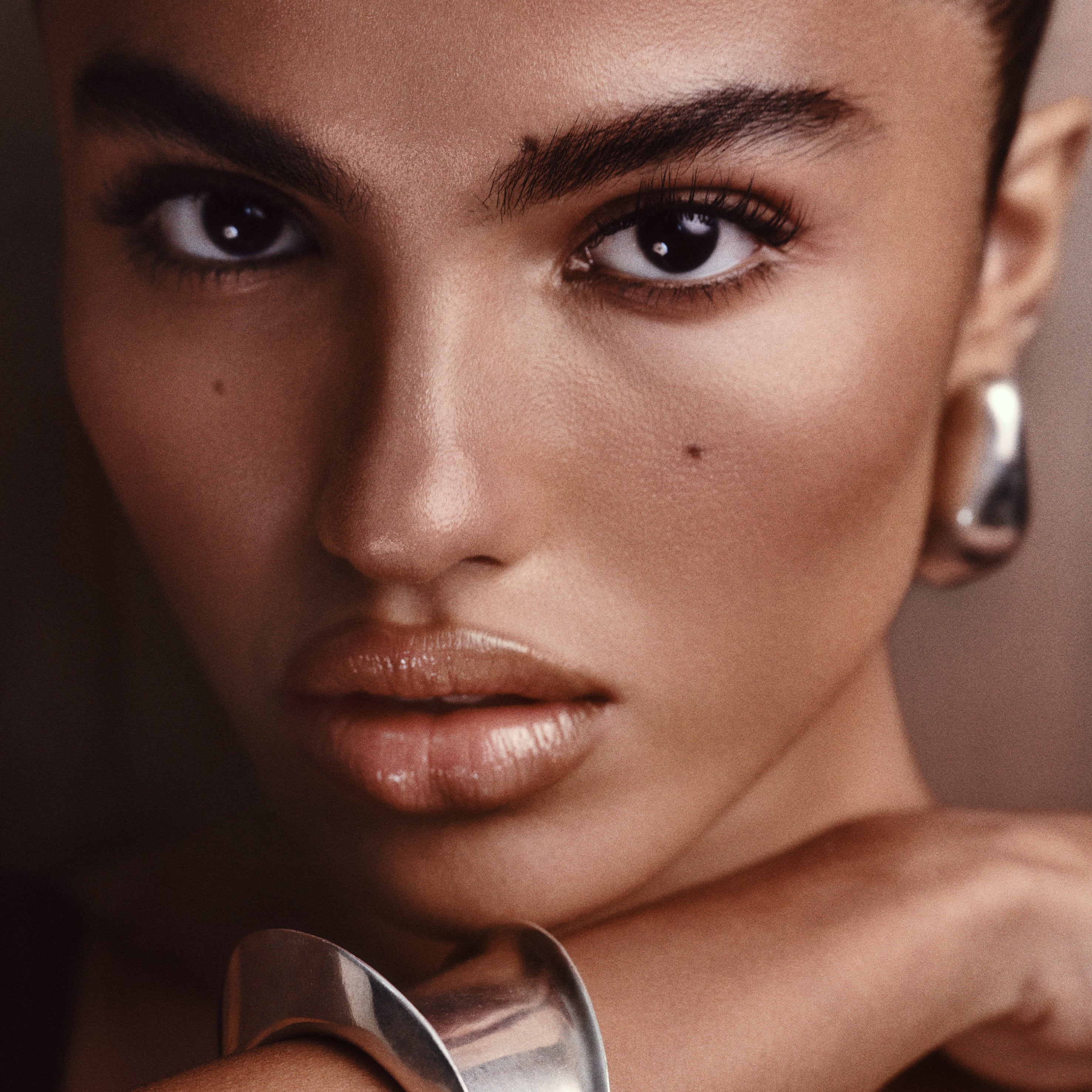 Close-up portrait of a model wearing a chunky silver cuff bracelet and matching silver hoop earring against a warm brown background.