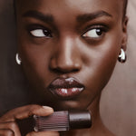 Close-up portrait of a woman holding a ribbed dark lipstick tube, with glossy mauve-brown lipstick on her lips against a soft taupe background.