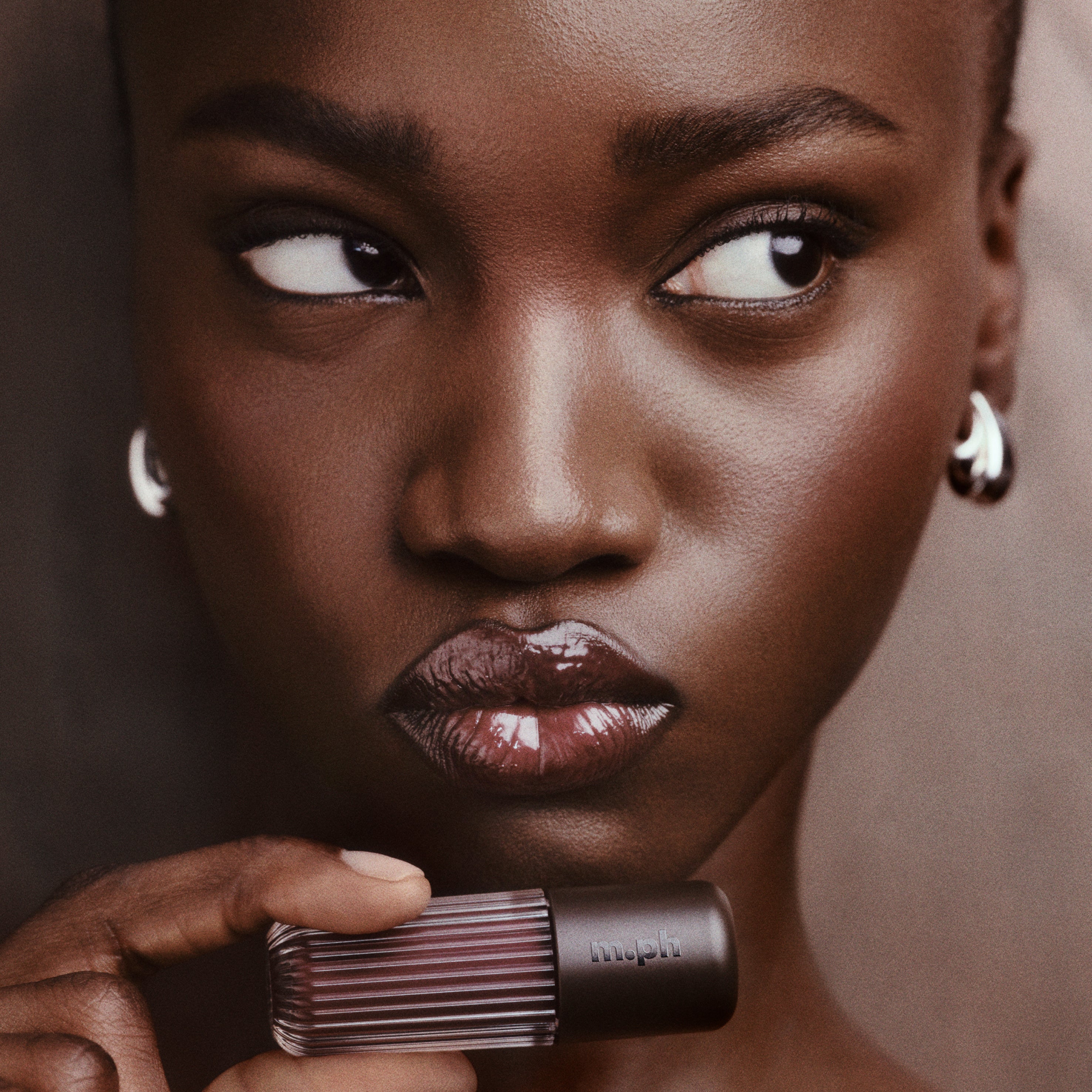 Close-up portrait of a woman holding a ribbed dark lipstick tube, with glossy mauve-brown lipstick on her lips against a soft taupe background.