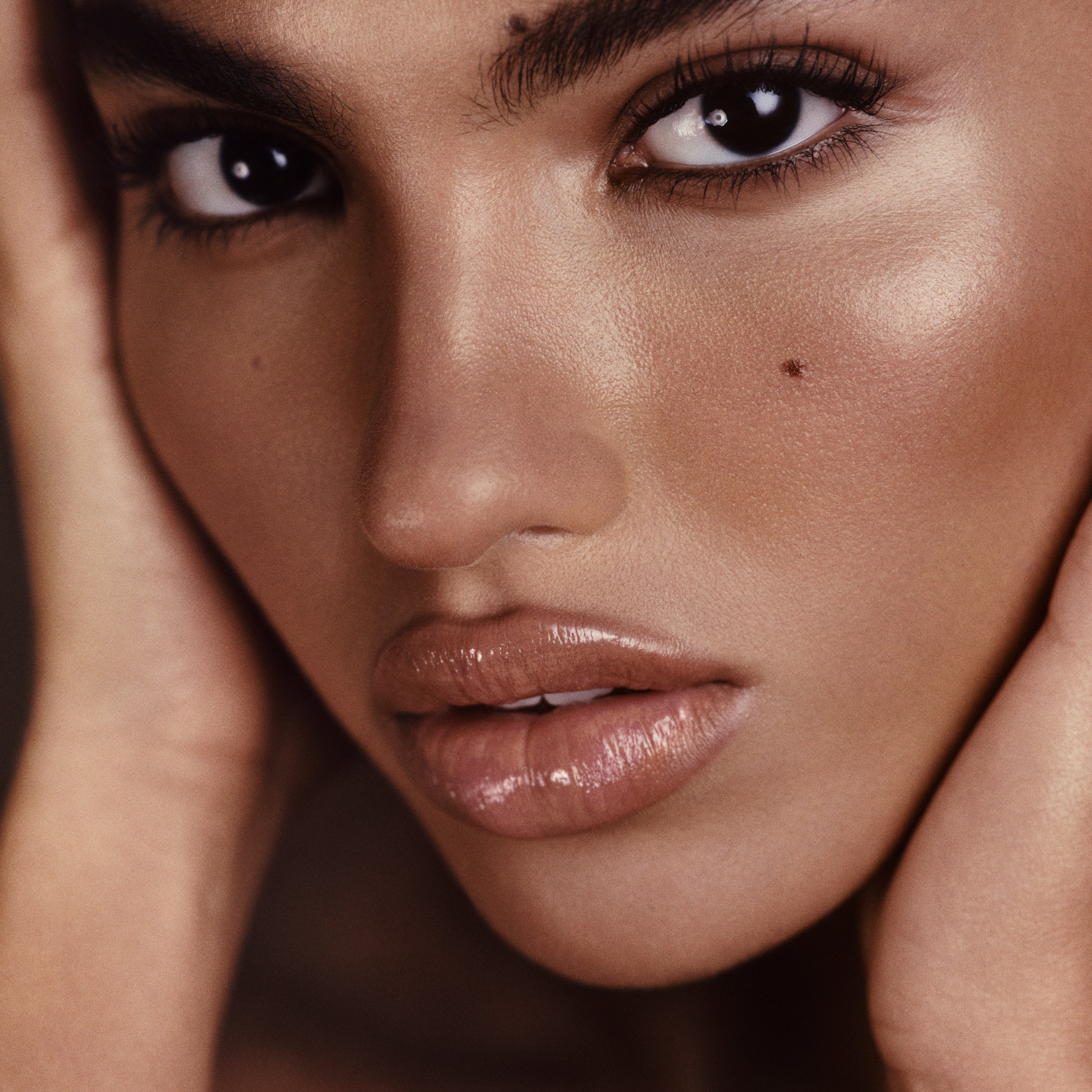 Close-up portrait of a woman with glossy nude lips, warm bronzed skin, and dark brown eyes, framed by her hands against a soft warm brown background.