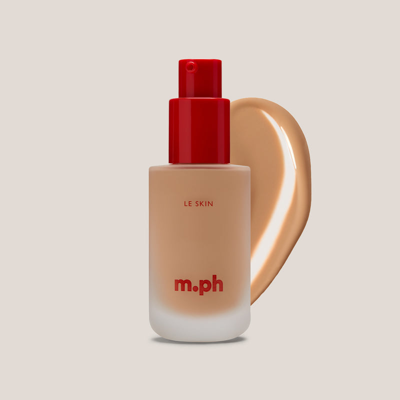 M.PH Le Skin sandy beige full coverage foundation in shade 100 with a silky texture swatch behind the frosted glass bottle.